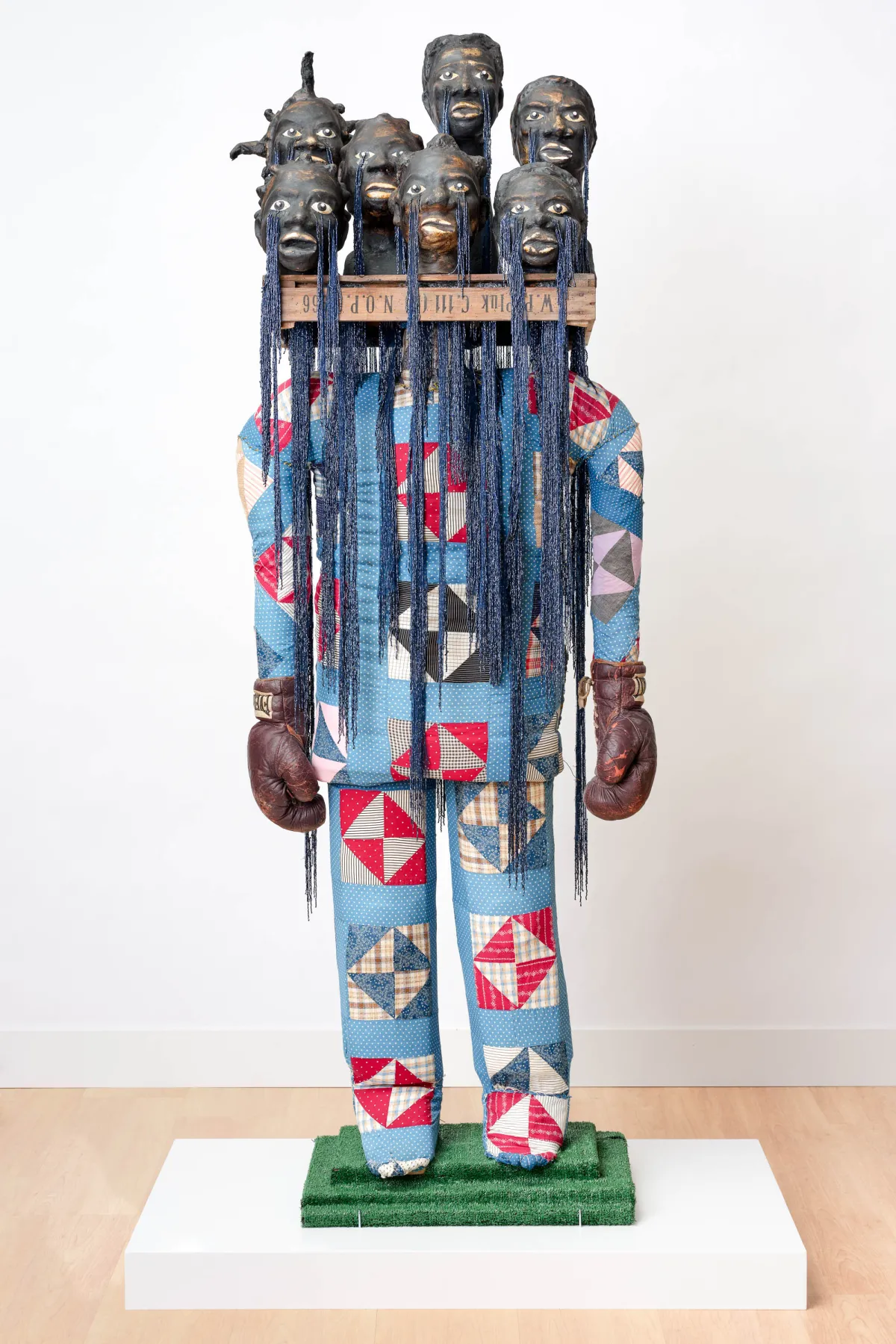 A standing figurative sculpture, approximately life-size, with a body made entirely of a quilted fabric jumpsuit in blue, red, and white geometric patterns. The figure wears large brown boxing gloves. A wooden crate rests on its shoulders, supporting a cluster of nine dark-skinned carved wooden heads with distressed, open-mouthed expressions and long, blue, beaded hair that covers the figure's chest.
