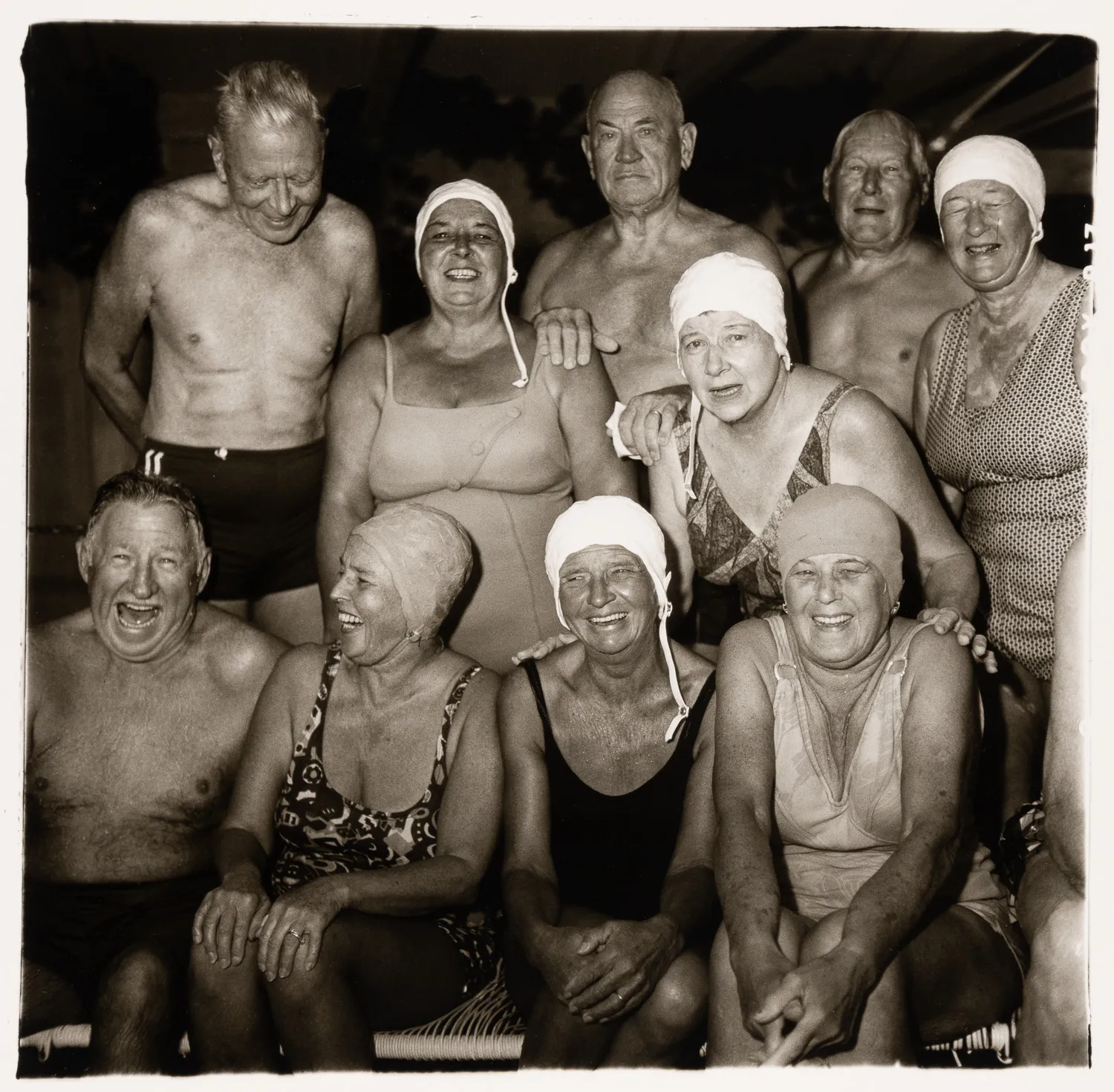A black-and-white group portrait of ten older adults—men and women—posing together. All the women are wearing white swim caps and modest swimsuits, and the men are shirtless in swim trunks. Many of them are laughing or smiling broadly at the camera.