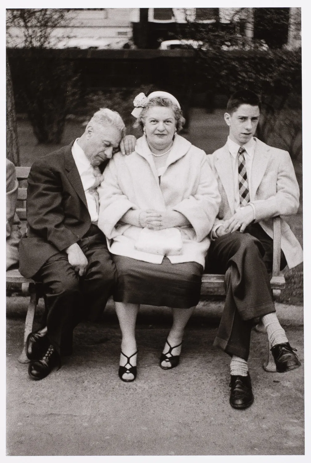 A black-and-white photo of an elderly couple and a young man sitting side-by-side on a park bench. The father, in a dark suit, leans his head asleep on the mother's shoulder. The mother, wearing a light coat and pearl necklace, sits straight-backed and looks at the camera. The young man, in a light blazer and tie, sits stiffly to the right.