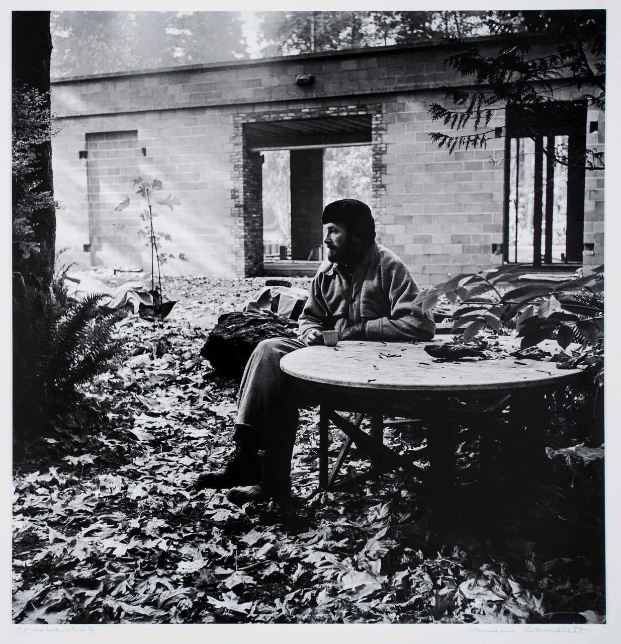 A black and white photograph of a man sitting outdoors at a rustic wooden table, surrounded by fallen leaves. He wears a heavy jacket and a beanie, holding a cup in one hand while gazing into the distance. Behind him is a partially constructed or abandoned concrete block building with open doorways and exposed beams. The surrounding environment is wooded, with ferns and trees framing the scene, evoking a quiet, contemplative atmosphere. The image is dated "October 1949" in the lower left corner.