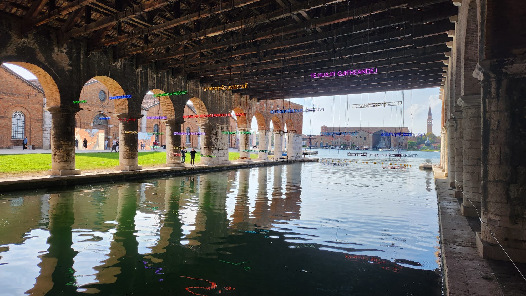 A covered port with brick archways shows neon signs by Claire Fontaine saying Foreigners Everywhere in different languages in in Venice Italy