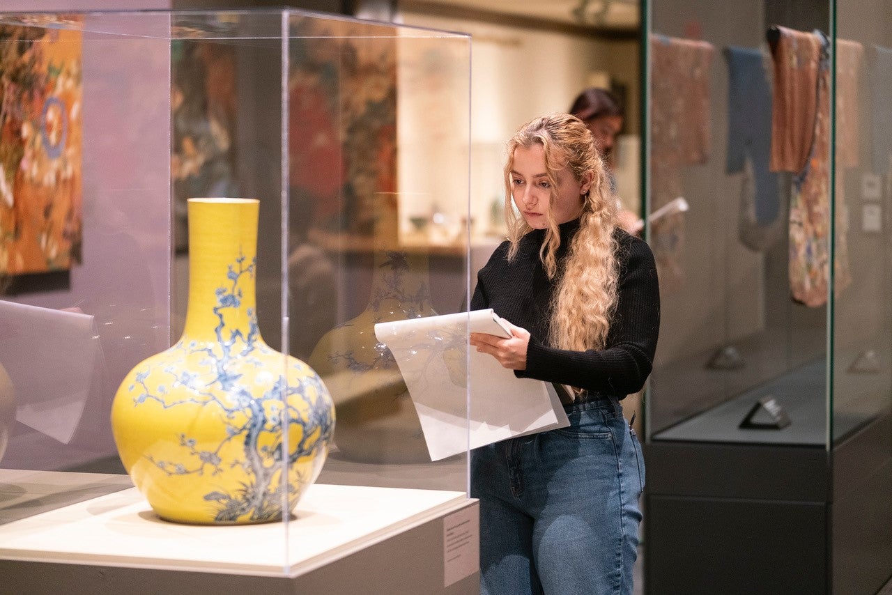 Yellow vase with blue flowers is on a stand in a museum. Blond girl with curly hair draws the vase.