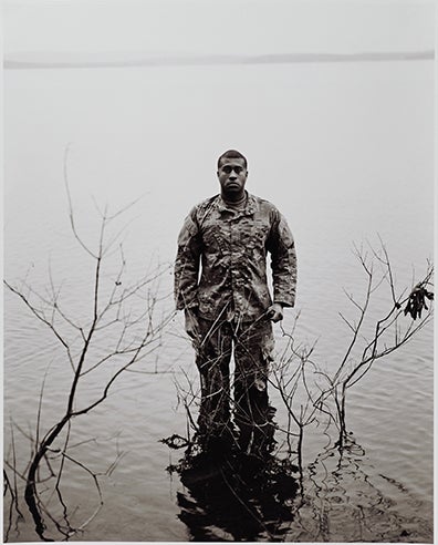 A soldier in camo standing knee deep in a lake with dead branches around him.
