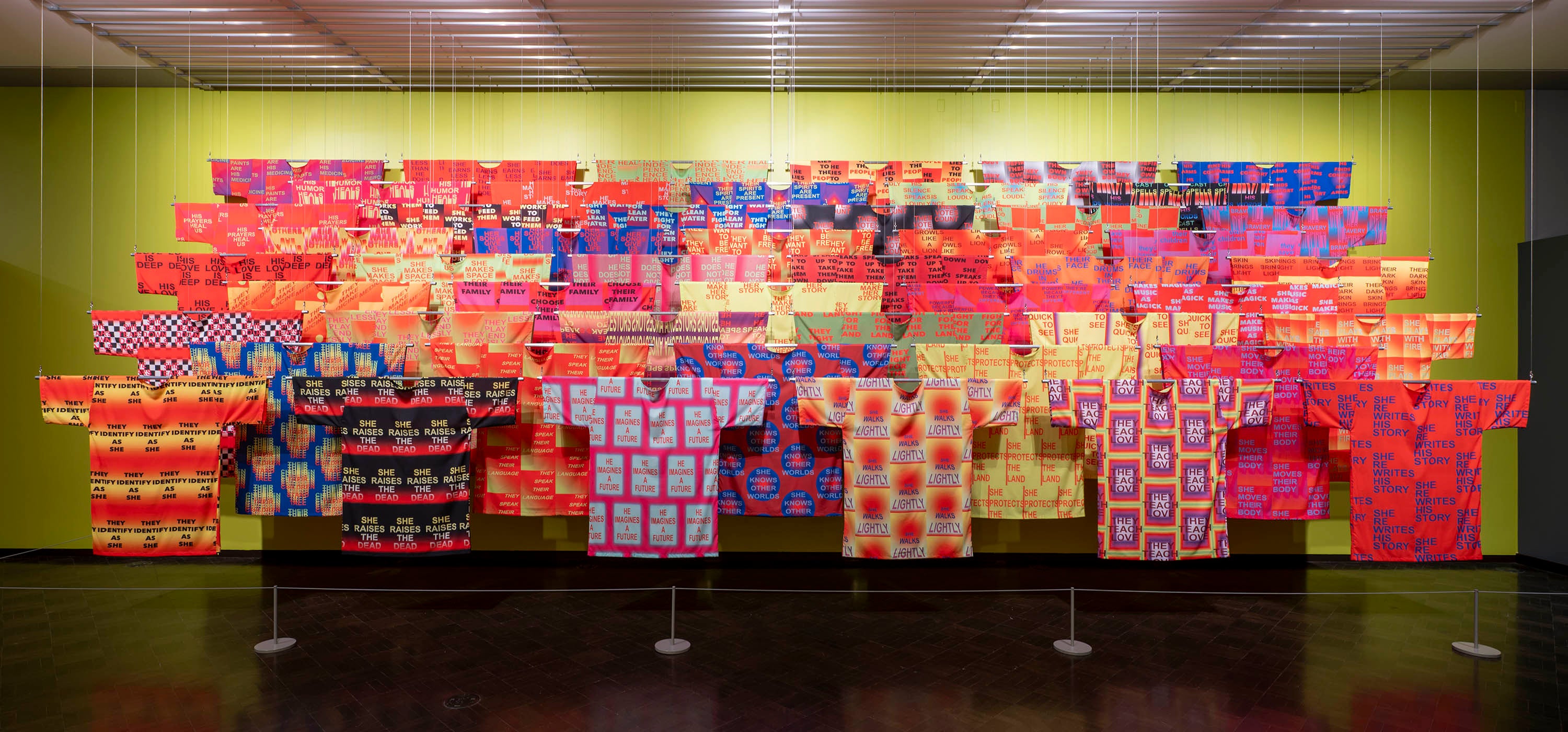 An art installation of many hanging shirts in rows, very colorful with geometric designs and words printed on them