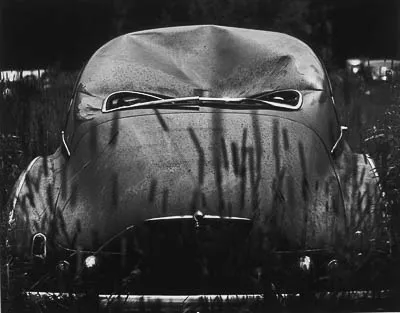 Black and white photograph of an old car with a dented hood and headlights that appear to peer through tall grass.