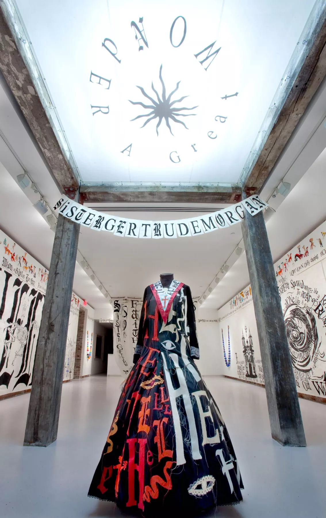 An installation art piece featuring a black dress adorned with colorful words and symbols, displayed in a gallery setting. Above the dress, a banner reads "SISTER GERTRUDE MORGAN," and the walls are covered with related text and imagery.