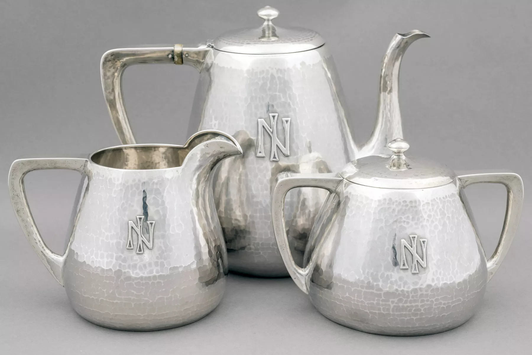 Three silver pitchers are arranged artfully on a grey background. The same symbol which looks like an N with a line vertically through the middle are on all three.