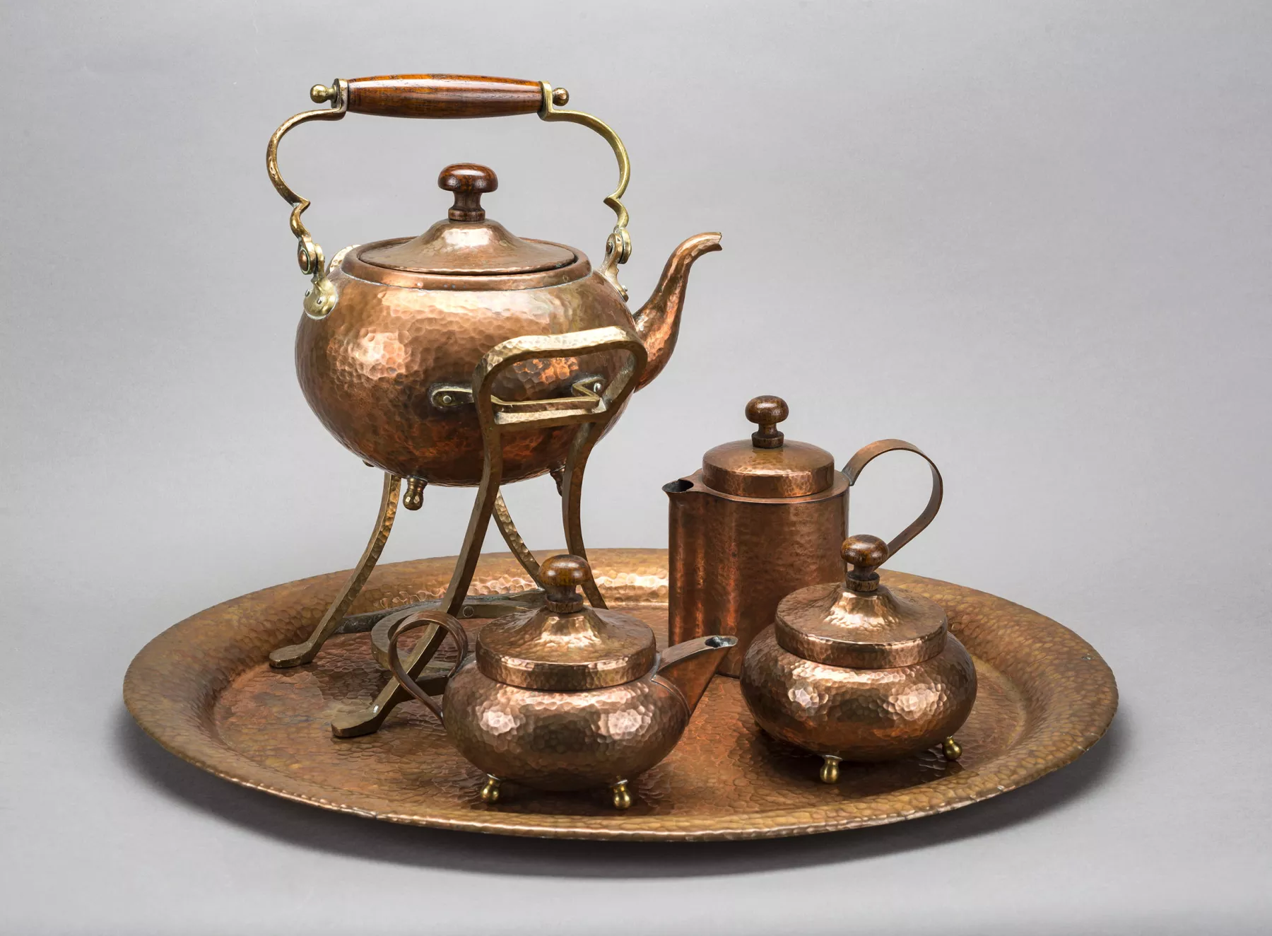 A copper serving tray with four tea service pitchers on it. The copper is hammered and dimpled and they are placed on a grey backdrop.