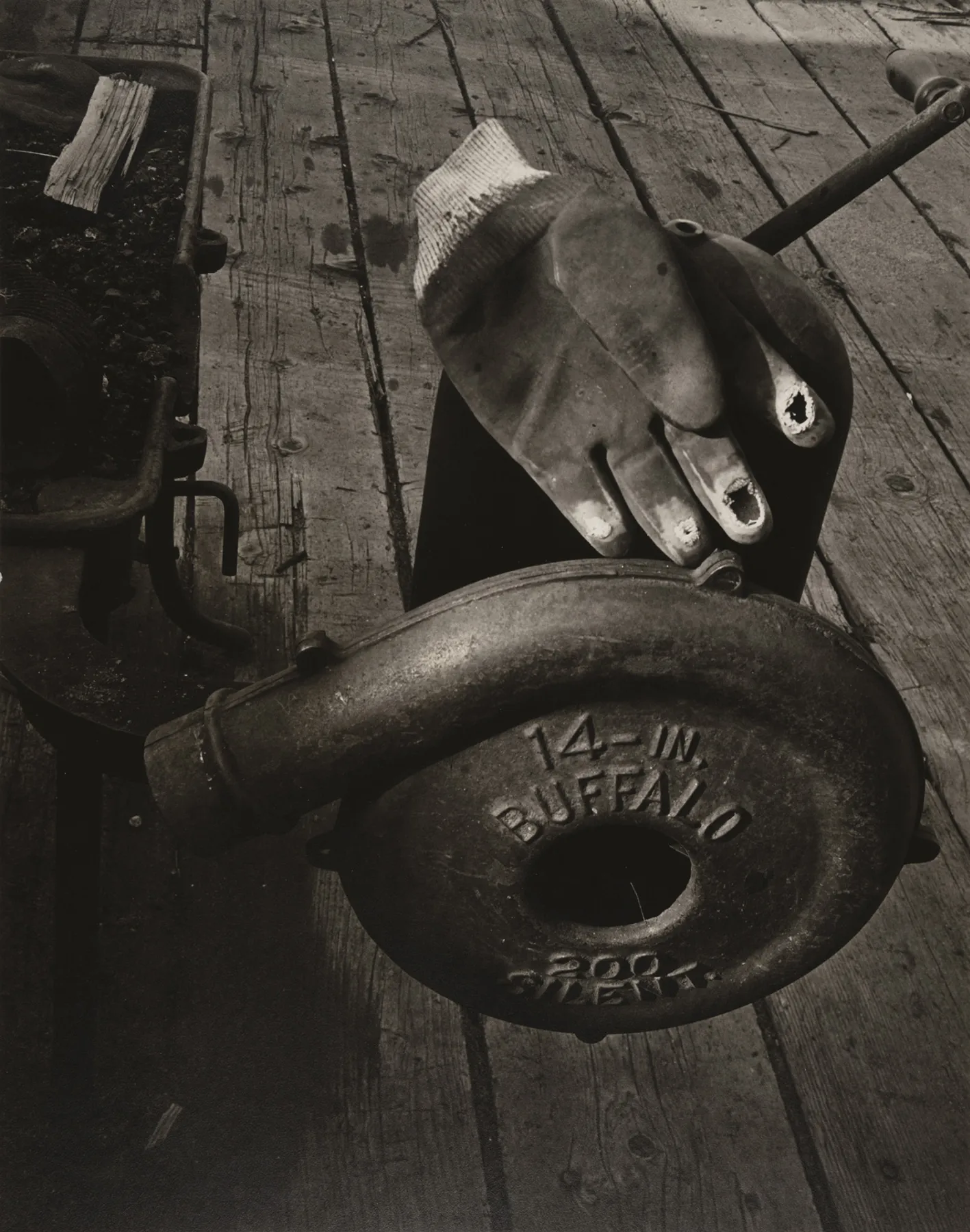 A black and white photograph featuring a worn work glove with holes in the fingertips resting on a piece of industrial equipment. The equipment has a circular metal casing embossed with the words "14-IN. BUFFALO 200 SILENT." The setting is a wooden work surface with visible cracks and stains. To the left, a metal cart filled with debris and a stack of papers or rags is partially visible. The composition highlights the textures of the glove, metal, and wood, evoking themes of labor and industrial history.