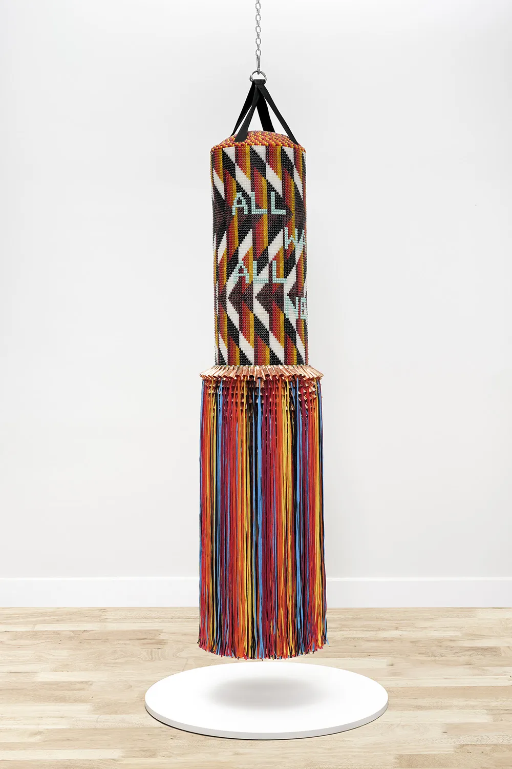 A sculptural work resembling a heavy punching bag suspended by a chain in a gallery. The top half is encased in intricate beadwork with a geometric pattern of black, white, red, and yellow zigzags. Light blue beaded text on the bag reads "ALL WAYS ALL NOW." A row of copper-colored metallic cones separates the beaded section from a long, dense skirt of multicolored fringe (red, blue, yellow, and black) that reaches toward a white circular base on the floor.