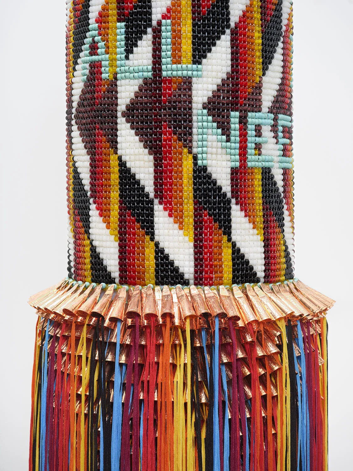 A close-up detail of the "heavy bag" sculpture. The image shows the fine craftsmanship of the geometric beadwork in black, white, red, and yellow, with a portion of the light blue beaded text visible. Below the beads, a dense row of copper-colored metallic cones is attached, with long strands of vibrant blue, red, yellow, and black fringe hanging from them.